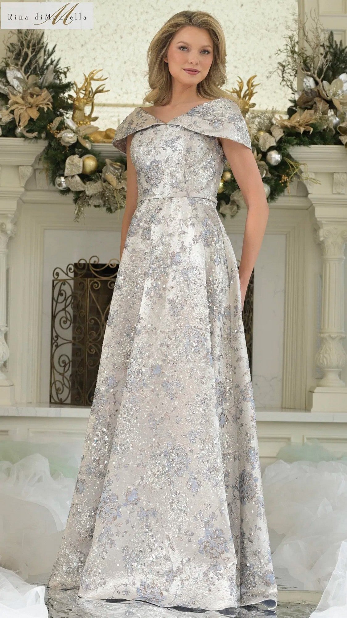 A woman stands indoors in front of a white ornate fireplace decorated with gold and white flowers, wearing the Rina Di Montella RD3312 long formal dress in Blue Multi.